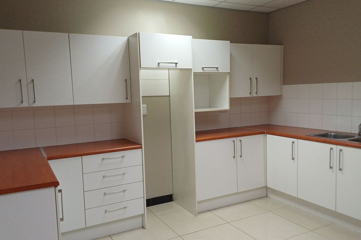 A clean, functional office kitchenette featuring white cabinetry, laminate countertops, a stainless steel sink, and tiled backsplash.