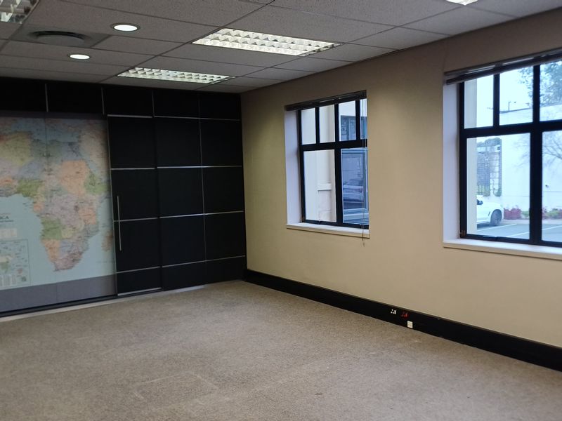A vacant office space featuring neutral carpeting, a suspended ceiling with recessed lighting, and dark feature wall paneling with large windows.