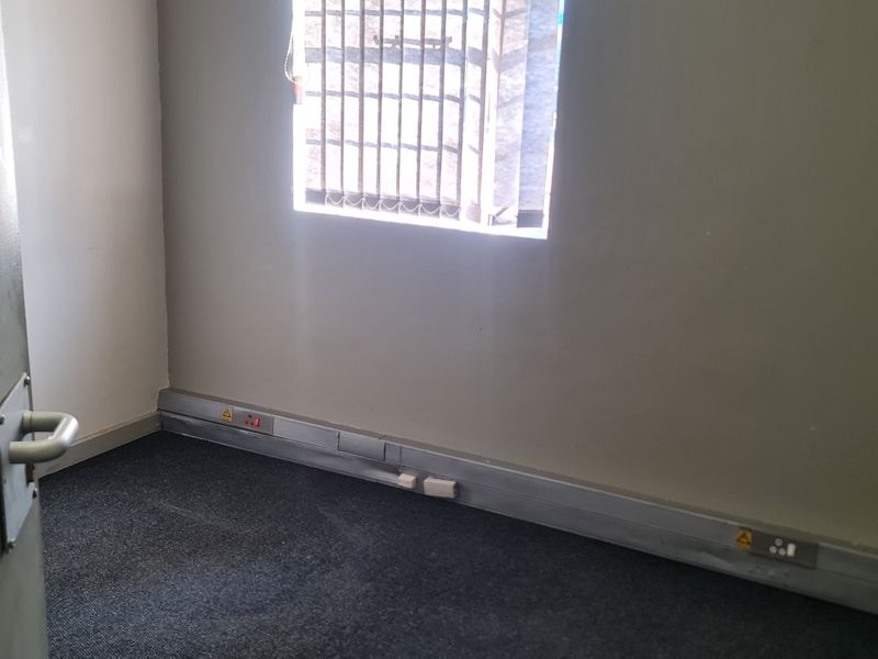 A small, empty office space featuring carpeted flooring, surface-mounted electrical trunking, and a window fitted with security bars.