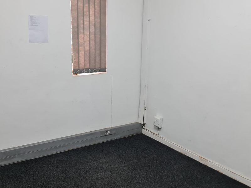 A small, empty office space featuring dark carpet flooring, vertical window blinds, and wall-mounted electrical outlets, requiring minor cosmetic repairs.
