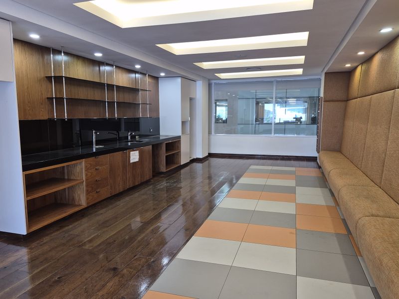 A modern office kitchen area featuring sleek wooden cabinetry, integrated sinks, recessed ceiling lighting, and a comfortable upholstered bench seating area.