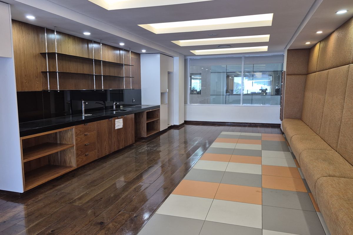 A modern office kitchen area featuring sleek wooden cabinetry, integrated sinks, recessed ceiling lighting, and a comfortable upholstered bench seating area.