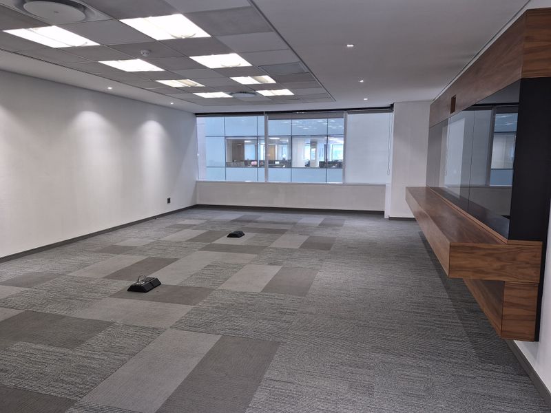 A spacious, empty office area featuring modern carpet tiles, a suspended ceiling with recessed lighting, and a sleek built-in wooden cabinetry unit.