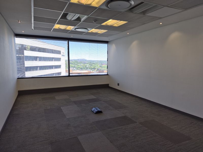 A vacant, well-lit office space featuring carpet tiles, a suspended ceiling with recessed lighting, and large windows offering an urban view.