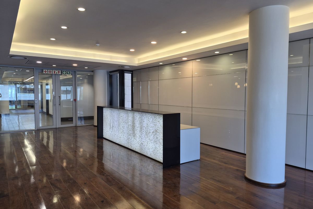Modern office reception area featuring polished hardwood flooring, a backlit reception desk, integrated wall cabinetry, and professional recessed lighting.