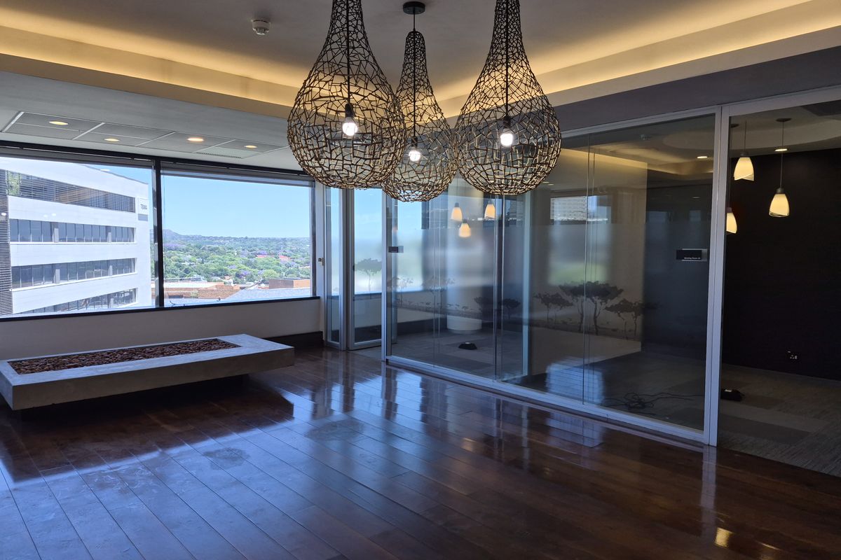 Modern office space featuring polished hardwood floors, glass partitions, and large windows with expansive city views, complemented by contemporary pendant lighting.