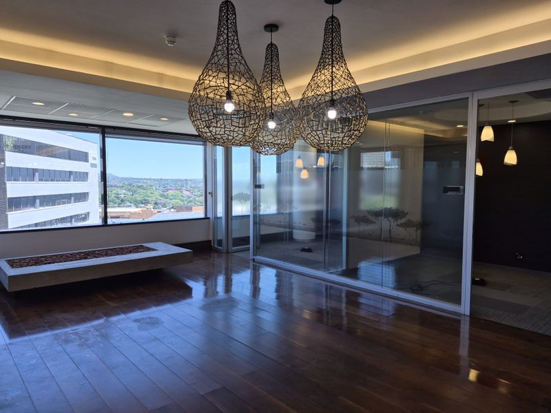 Modern office space featuring polished hardwood floors, glass partitions, and large windows with expansive city views, complemented by contemporary pendant lighting.
