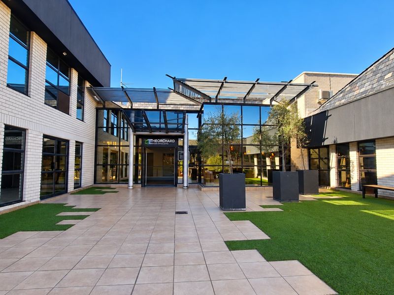 Modern office building entrance featuring a paved courtyard, artificial turf, and a glass-fronted facade with a metal pergola structure.