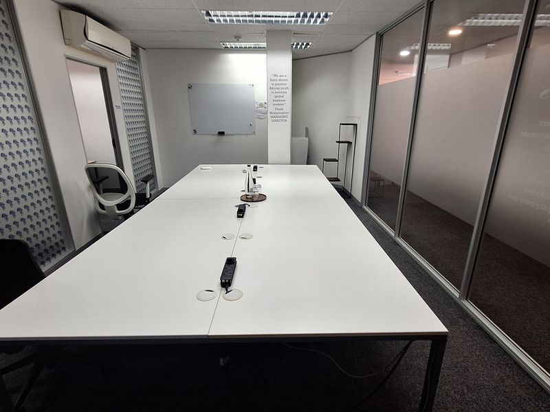 A modern office meeting room featuring a large central desk, glass partition walls, recessed ceiling lighting, and a wall-mounted whiteboard.