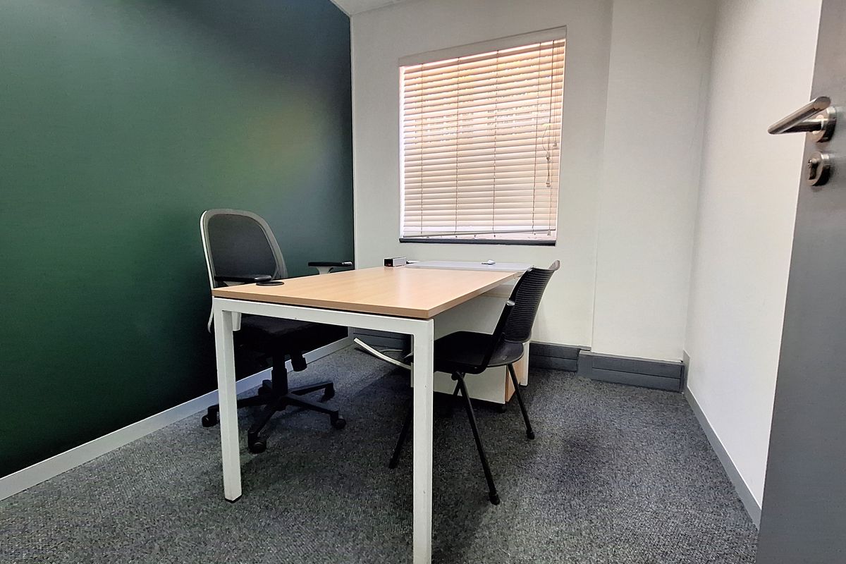 A compact office space featuring a dark green accent wall, carpeted flooring, and a window fitted with venetian blinds.