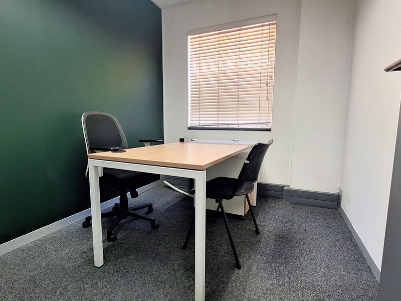 A compact office space featuring a dark green accent wall, carpeted flooring, and a window fitted with venetian blinds.