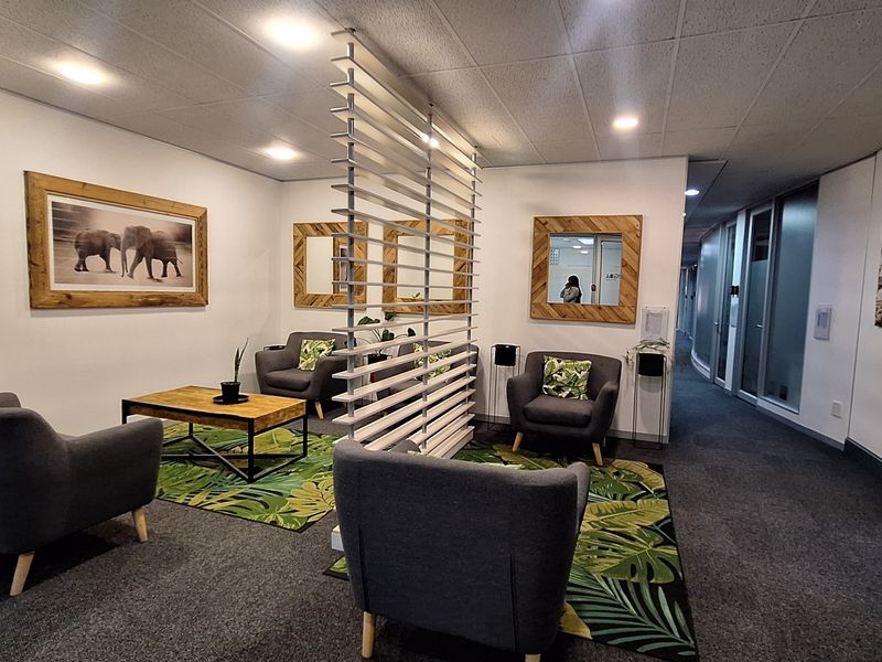 A modern office reception or breakout area featuring a slatted room divider, recessed ceiling lighting, and glass-partitioned office corridors.