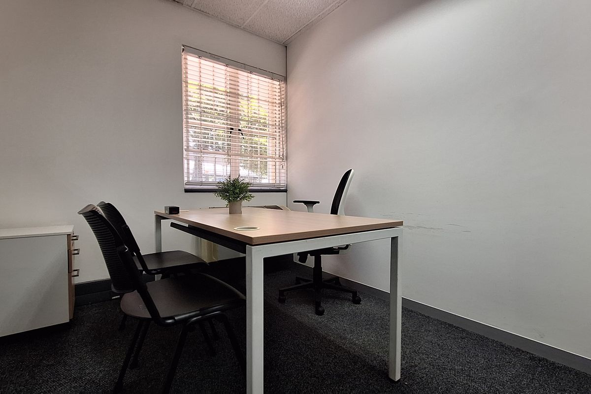 A compact office space featuring carpeted flooring, a suspended ceiling, and a window with venetian blinds, providing a functional workspace environment.
