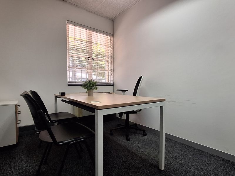 A compact office space featuring carpeted flooring, a suspended ceiling, and a window with venetian blinds, providing a functional workspace environment.