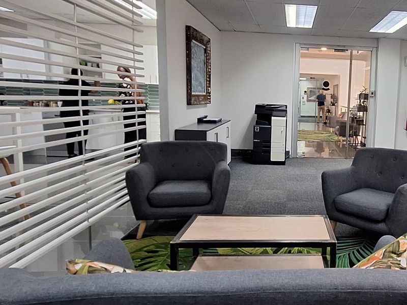 A professional office waiting area featuring carpeted flooring, a suspended ceiling with recessed lighting, and a glass partition separating the space from the kitchen.