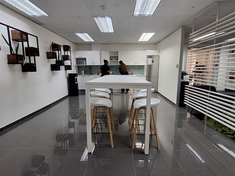 A modern office kitchen area featuring sleek white cabinetry, a tiled backsplash, recessed lighting, and polished floor tiles, providing a clean and functional break space.