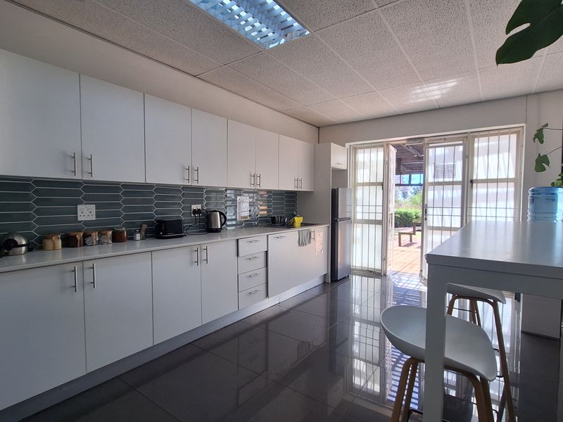 A modern office kitchen featuring white cabinetry, a dark tiled backsplash, polished floor tiles, and direct access to an outdoor patio area.