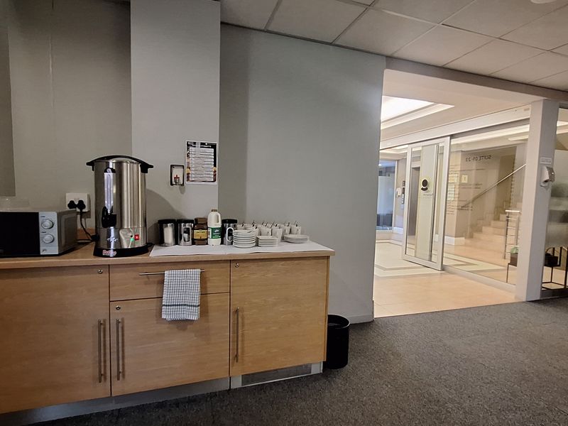 A functional office kitchenette area featuring built-in wooden cabinetry, carpeted flooring, and a glass partition leading to a bright lobby space.