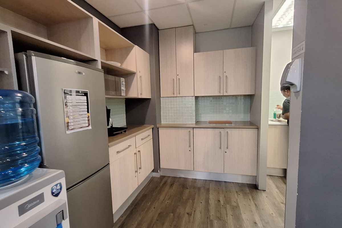 A functional office kitchenette featuring light wood-tone cabinetry, laminate countertops, a tiled backsplash, and durable laminate flooring.