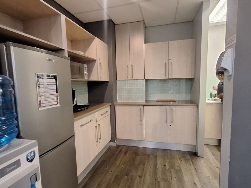 A functional office kitchenette featuring light wood-tone cabinetry, laminate countertops, a tiled backsplash, and durable laminate flooring.