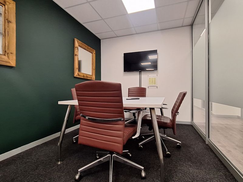 A modern office meeting room featuring a glass partition wall, dark green accent wall, carpeted flooring, and a wall-mounted television.