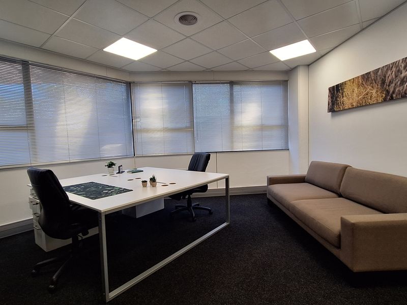 A professional office space featuring large windows with venetian blinds, recessed lighting, and dark carpeting, providing a clean and functional work environment.