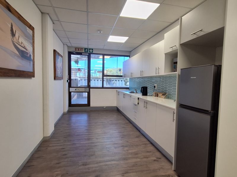 A clean and modern office kitchenette featuring white cabinetry, a tiled backsplash, laminate flooring, and a drop ceiling with recessed lighting.
