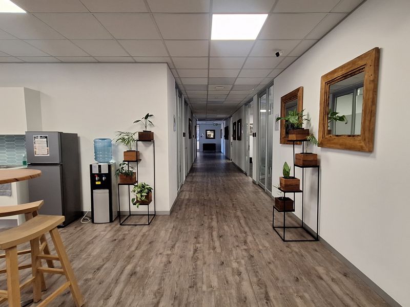 A modern office hallway featuring laminate flooring, suspended ceilings with recessed lighting, and glass-partitioned office spaces, adjacent to a small kitchenette area.