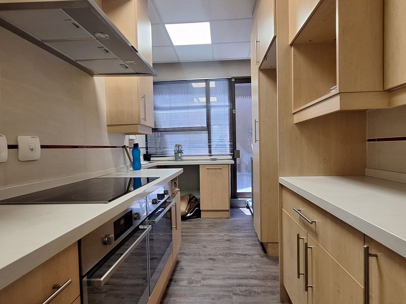 A clean, functional office kitchen featuring light wood cabinetry, integrated oven and hob, tiled backsplash, and modern laminate flooring.