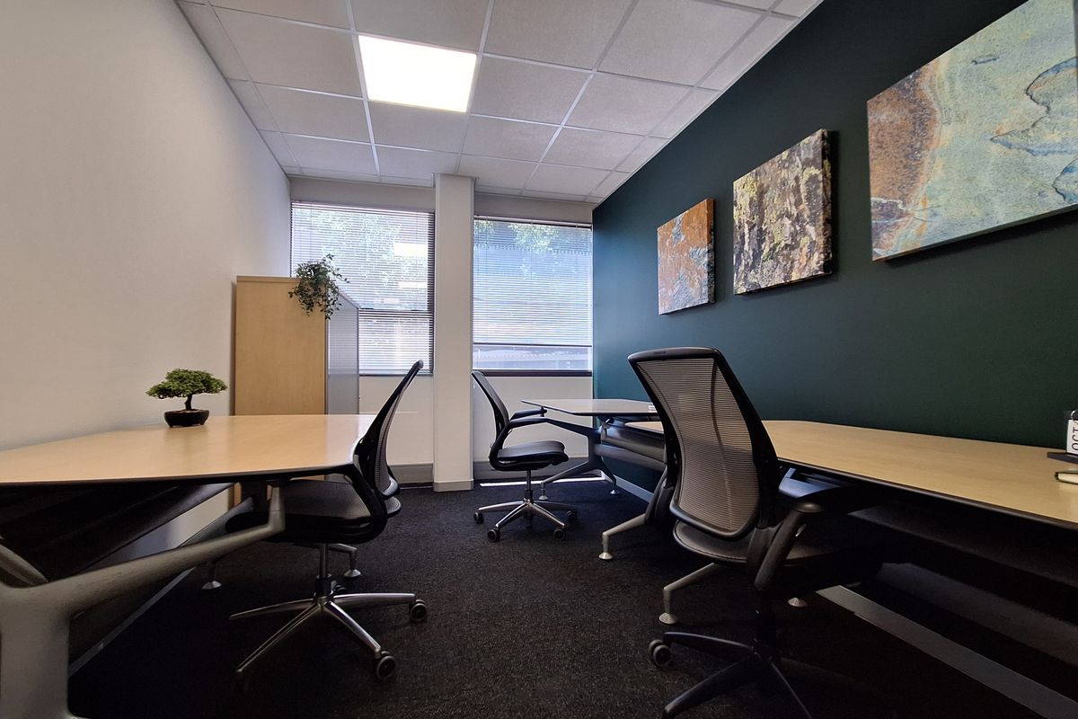 A modern, well-lit office space featuring carpeted flooring, a suspended ceiling with recessed lighting, and large windows with venetian blinds.
