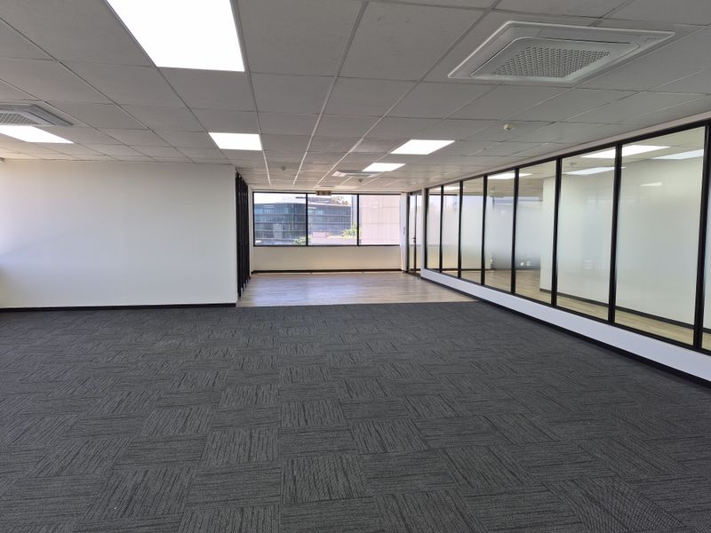 A spacious, empty open-plan office featuring modern carpet tiles, suspended ceilings with recessed lighting, and glass partition walls providing a bright, professional workspace.