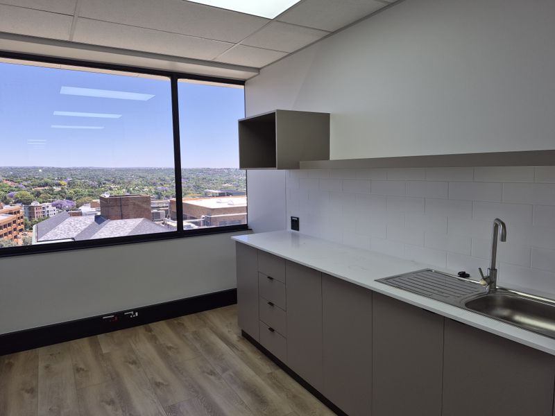 A modern, clean office kitchenette featuring sleek cabinetry, stone countertops, a stainless steel sink, and large windows offering an expansive city view.