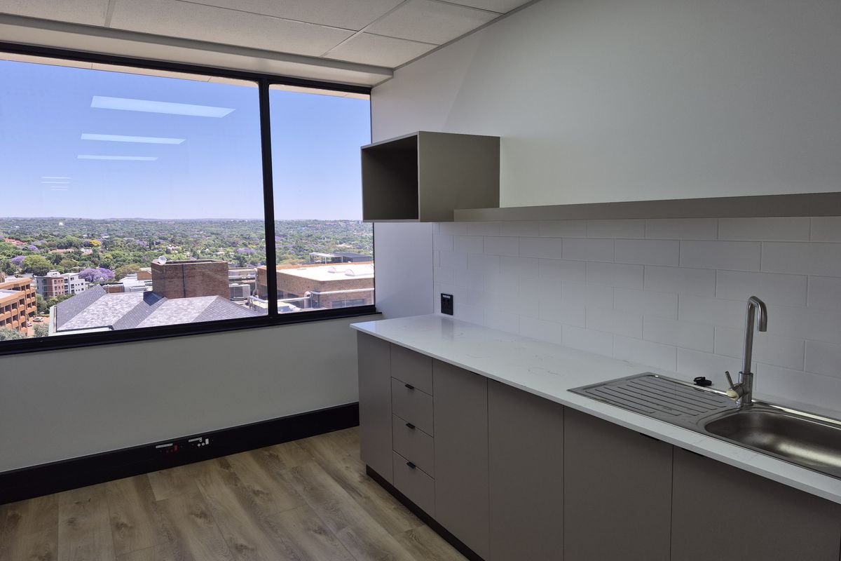 A modern, clean office kitchenette featuring sleek cabinetry, stone countertops, a stainless steel sink, and large windows offering an expansive city view.