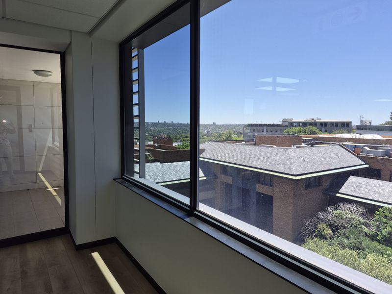 A bright, empty office space featuring large windows with expansive views, modern laminate flooring, and a glass partition wall.