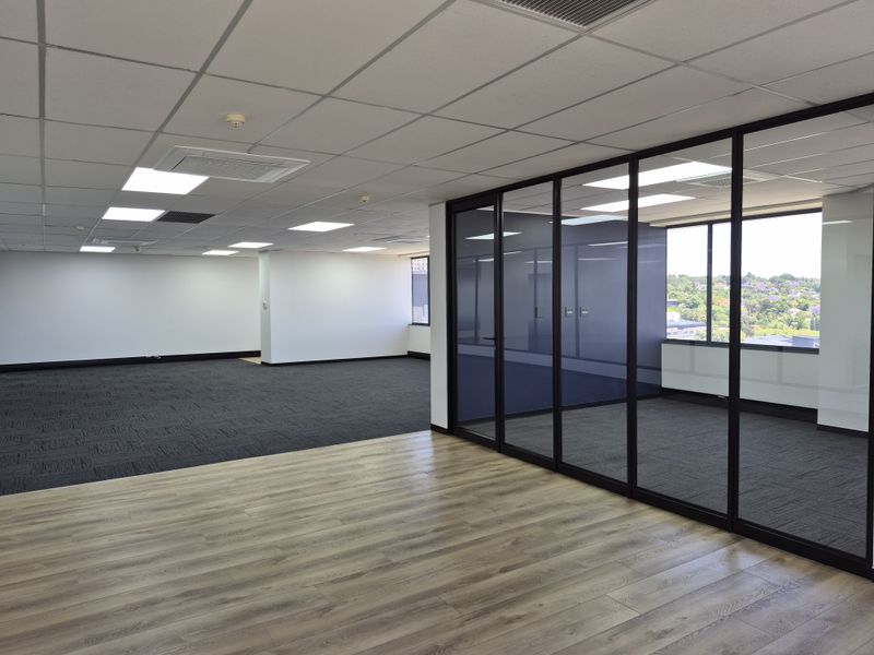Modern open-plan office space featuring a glass-partitioned meeting room, suspended ceiling, and a combination of carpet and wood-look laminate flooring.