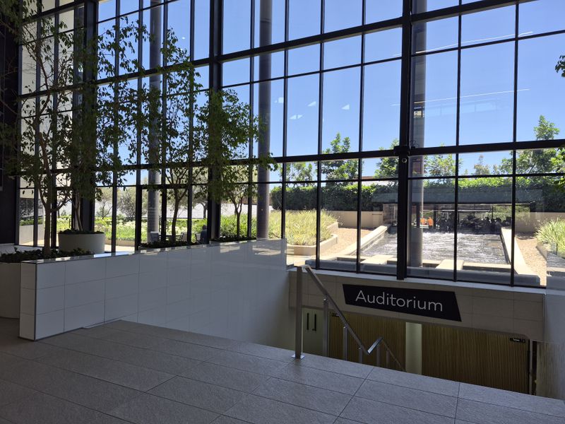 Modern office building entrance featuring a grand floor-to-ceiling glass facade, tiled flooring, and a staircase leading down to the auditorium.