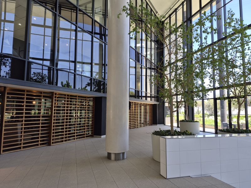A spacious, modern office building entrance featuring floor-to-ceiling glass walls, structural columns, and elegant timber slat detailing with integrated planters.