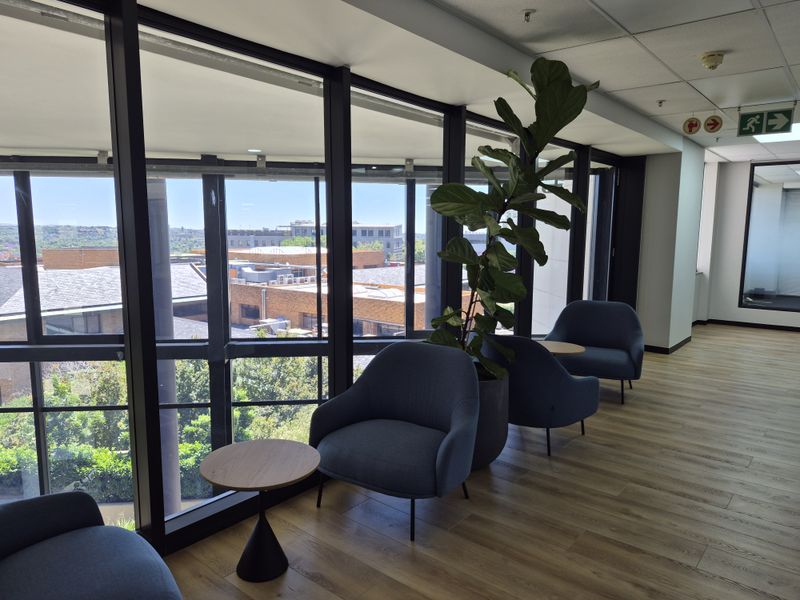 A bright office breakout area featuring floor-to-ceiling windows with exterior views, wood-look flooring, and integrated ceiling fixtures.