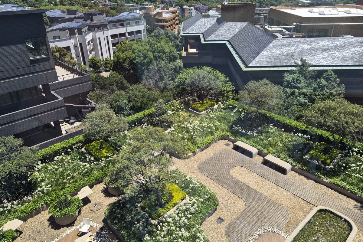 An elevated aerial view of a well-maintained rooftop garden featuring landscaped greenery, paved pathways, built-in stone seating, and a decorative water feature.