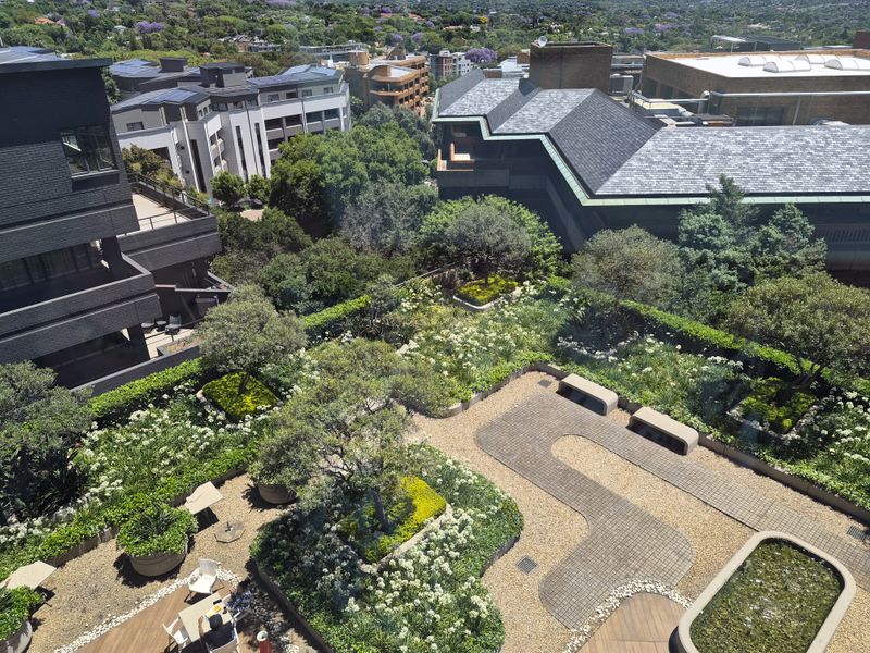 An elevated aerial view of a well-maintained rooftop garden featuring landscaped greenery, paved pathways, built-in stone seating, and a decorative water feature.