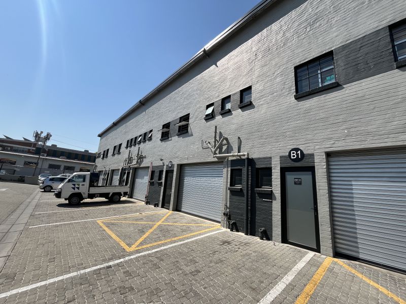 Exterior view of an industrial property featuring a grey brick facade, multiple roller shutter doors, and a paved parking area with designated bays.