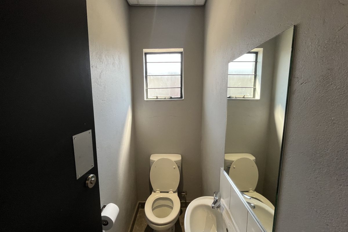A compact industrial-style bathroom featuring a toilet, wall-mounted basin, and a large wall mirror with a louvered window for ventilation.