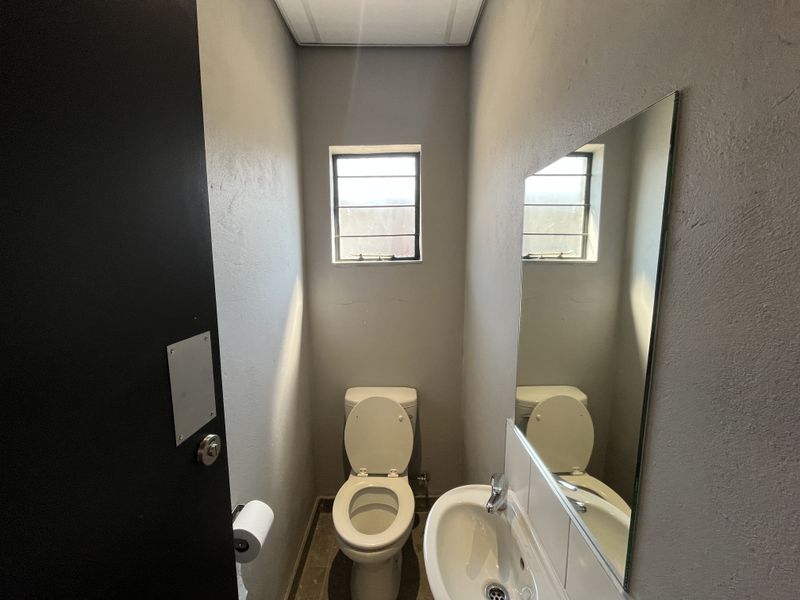 A compact industrial-style bathroom featuring a toilet, wall-mounted basin, and a large wall mirror with a louvered window for ventilation.