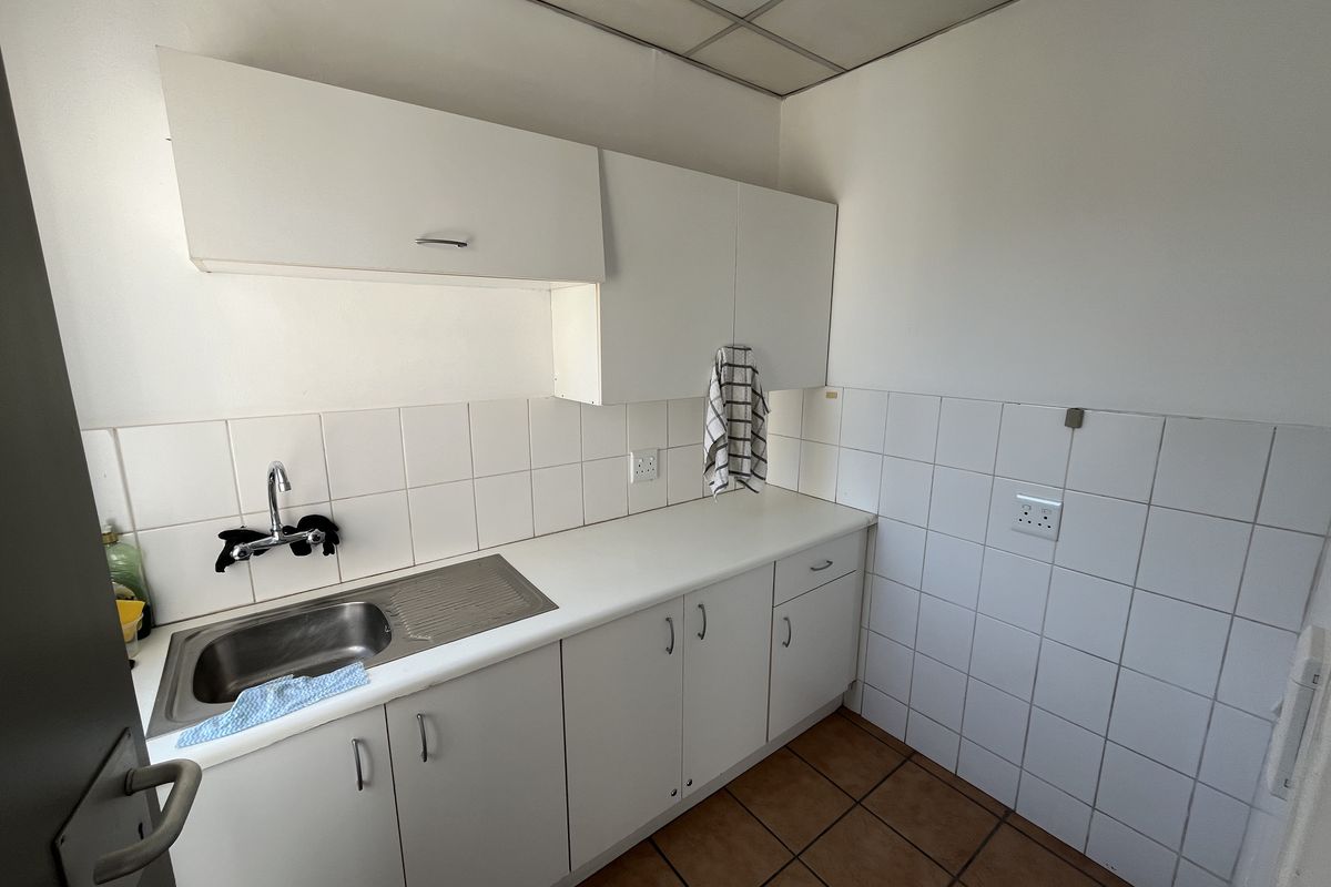 A functional kitchenette featuring white cabinetry, a stainless steel sink, tiled splashbacks, and tiled flooring, suitable for office use.