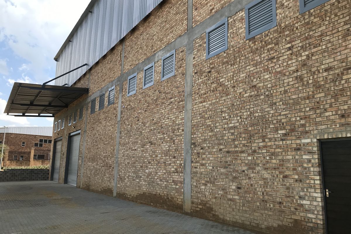 Side elevation of an industrial warehouse featuring face-brick walls, roller shutter doors, metal cladding, and ventilation louvres with paved exterior access.
