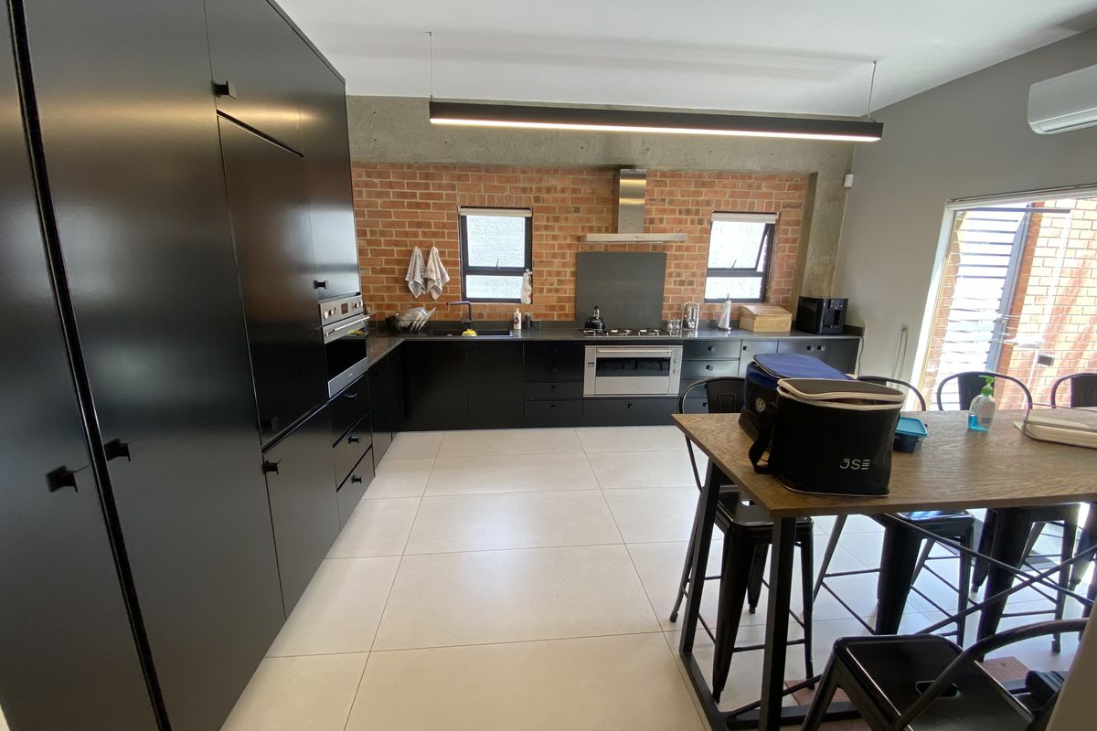Modern office kitchen featuring sleek black cabinetry, exposed brick wall, stainless steel appliances, and large format floor tiles.