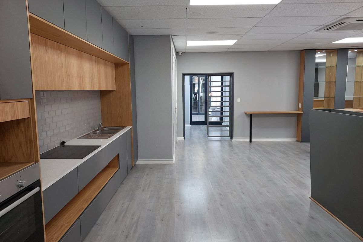 Modern office kitchenette featuring sleek grey cabinetry, wood-tone accents, integrated oven, hob, and a subway tile backsplash, leading to a secure glass-paneled entrance.