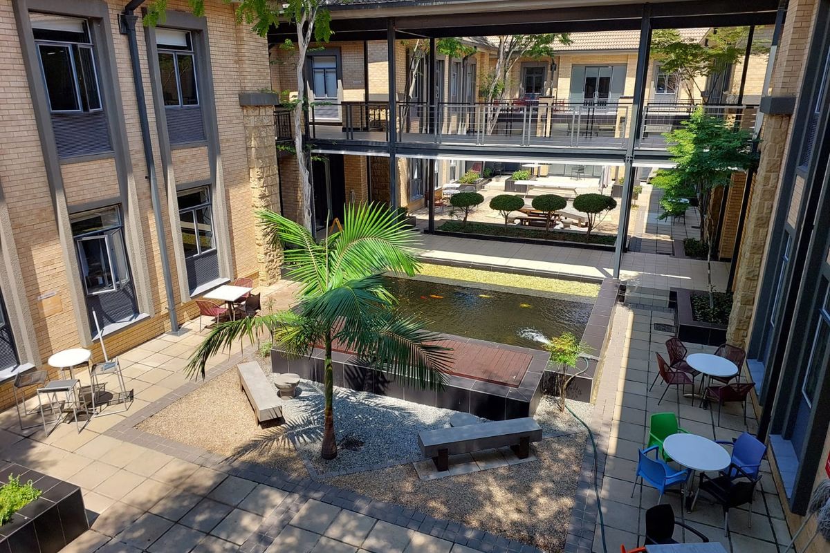 A well-maintained office courtyard featuring a central koi pond, paved seating areas, and a connecting overhead walkway between building wings.