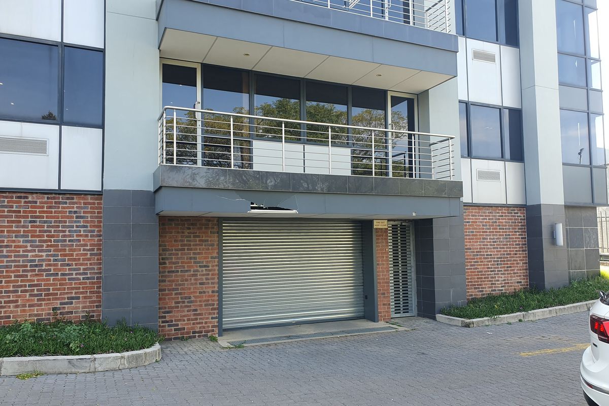 Exterior view of an office building featuring a roller shutter door, brick and panel facade, and a balcony with stainless steel railings.