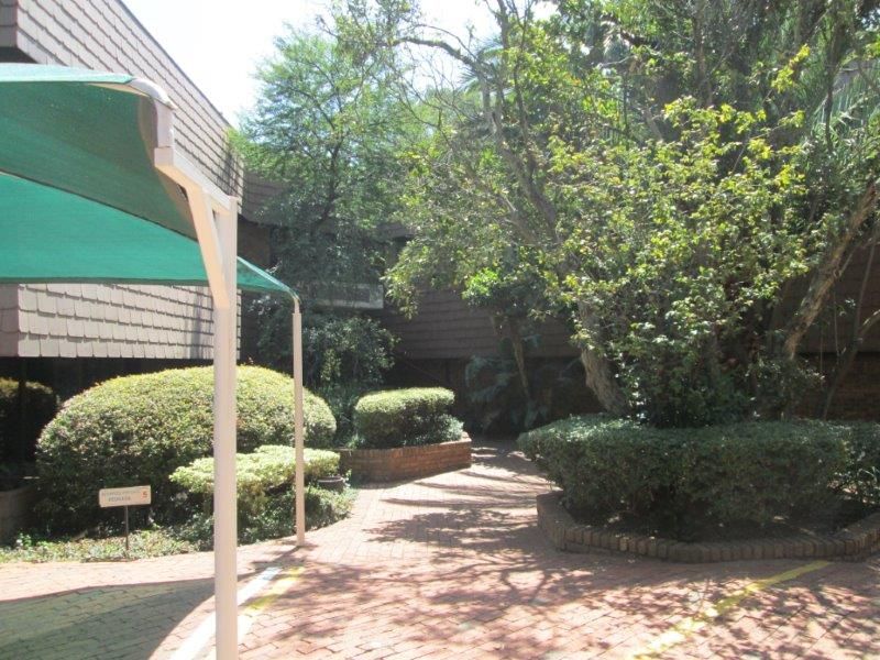 A landscaped outdoor area featuring brick paving, mature trees, established shrubbery, and a shaded parking or walkway structure.